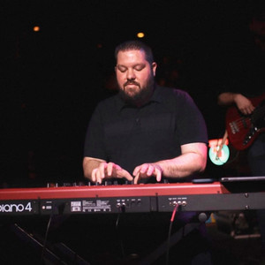 Man in a dark shirt playing an electronic keyboard on a dimly lit stage.